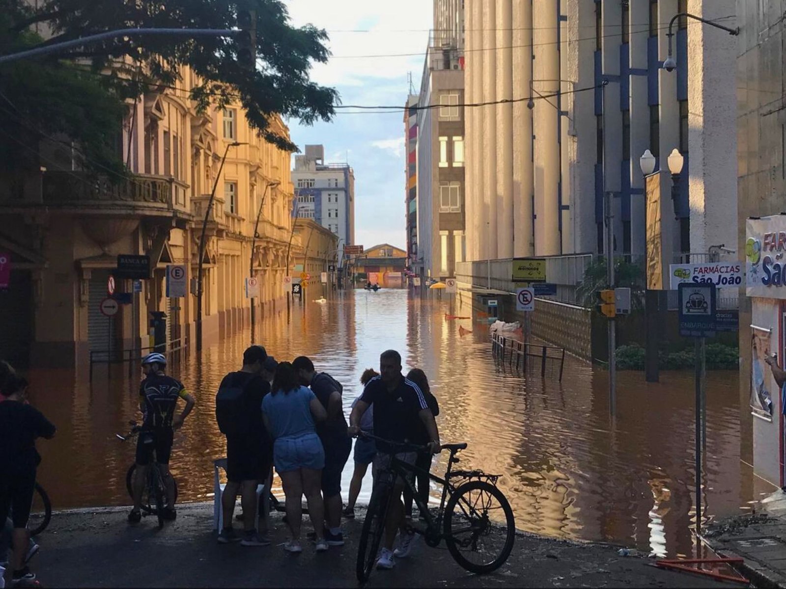 A imagem mostra enchentes em Porto Alegre, com a água invadindo o centro da cidade, enquanto cicloturistas observam a tragédia.