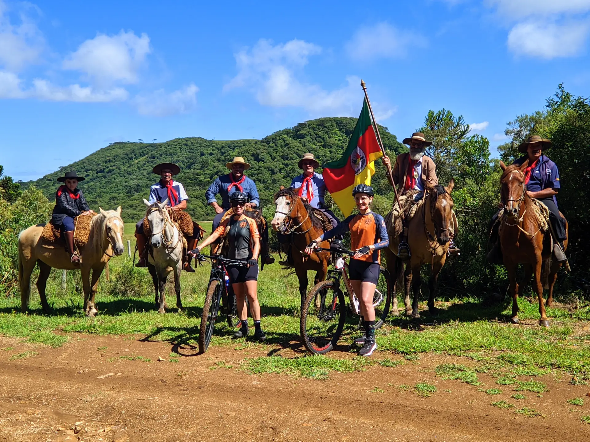 Grupo de tropeiros renascentes a cavalo e cicloturistas contemporâneos no Costão do Cambará, Campos de Cima da Serra (RS), com bandeira histórica e paisagem montanhosa. Foto: Ivan Mendes © Lobi Ciclotur