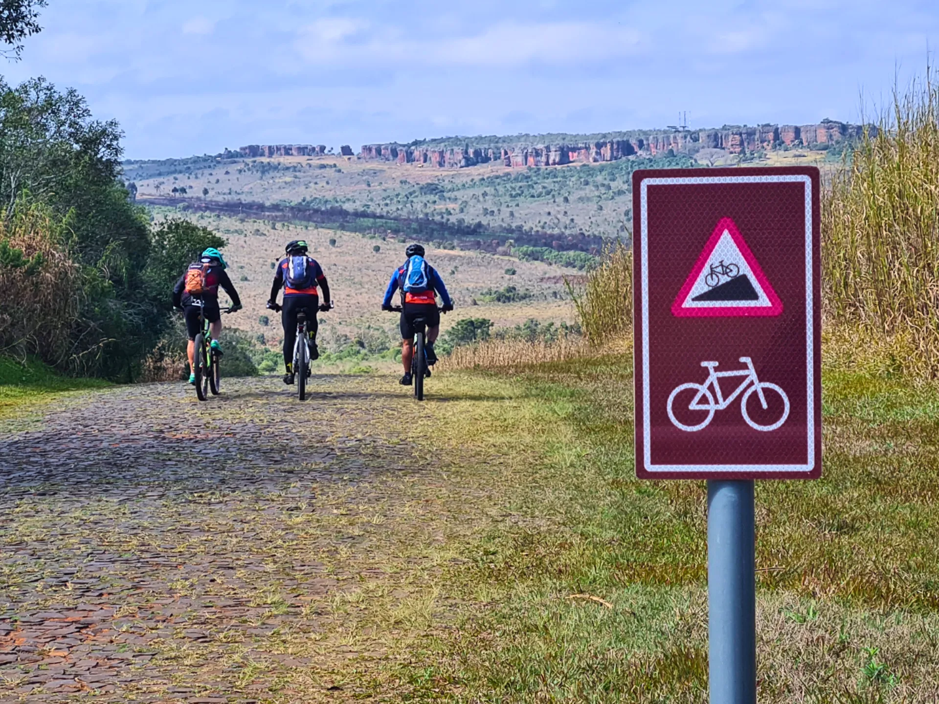 Placa de sinalização de cicloturismo no padrão Internacional no Parque Vila Velha, indicando aclive e rota de bicicletas com os Arenitos ao fundo.